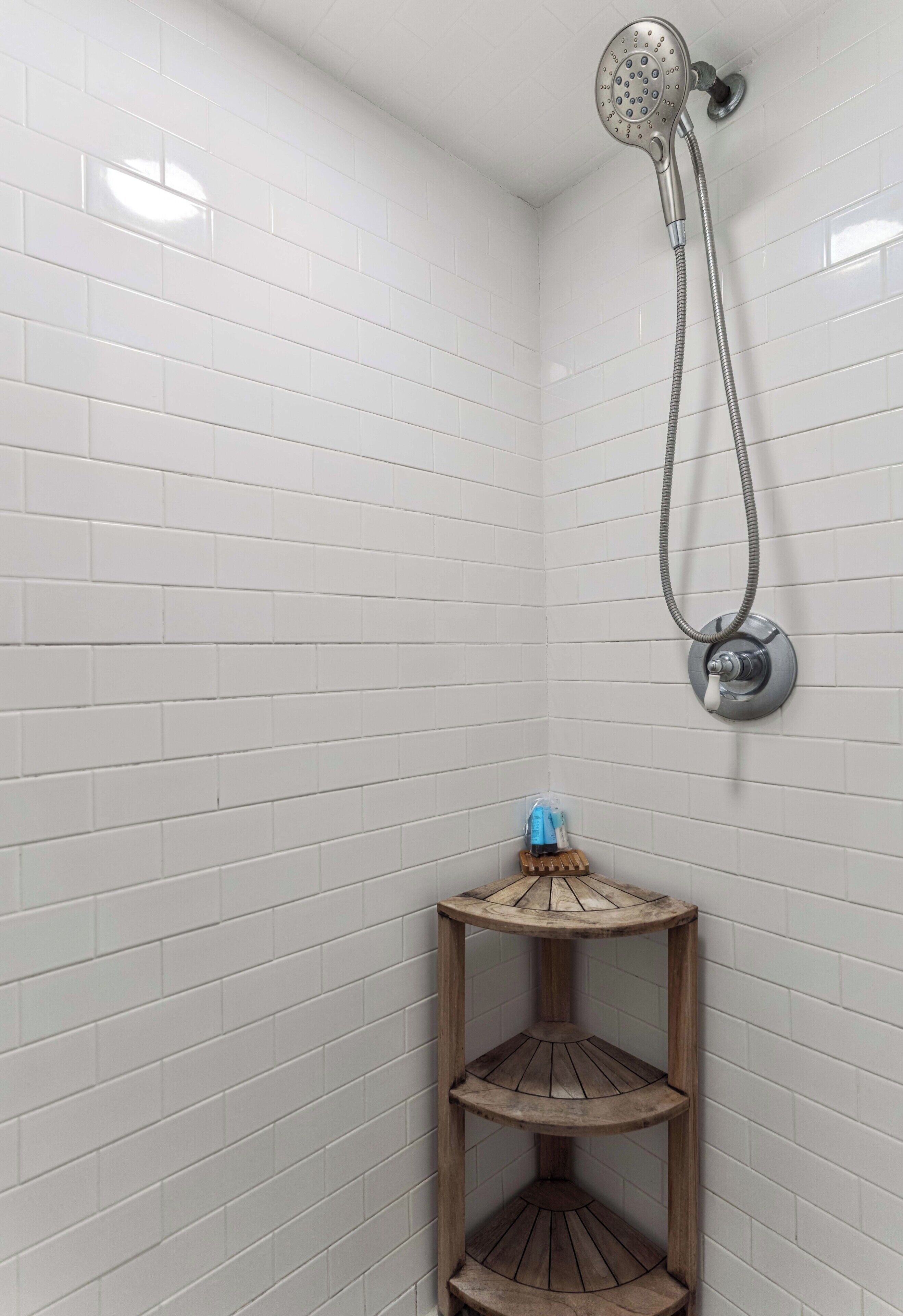 Zona de ducha con paredes de azulejos blancos, cabezal de ducha manual y un estante de madera con un objeto azul.