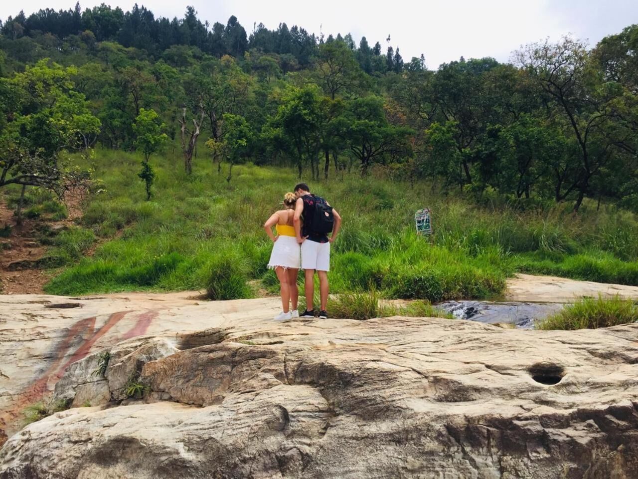 Two people standing on a rocky surface with a grassy area and trees in the background.