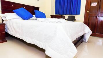 Un lit bien fait, avec une literie blanche et des oreillers bleus, une tête de lit en bois, une table de chevet avec une lampe, une chaise et une porte sur laquelle est apposé un panneau « interdiction de fumer ».