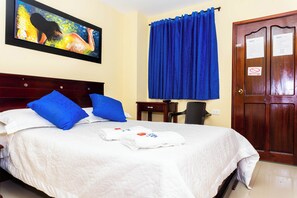 A neatly made bed with blue pillows, a painting on the wall, a blue curtain, and a wooden door with a sign. - Hotel Arce Plaza (Valledupar)