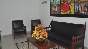 Een kamer met een zwarte bank, twee houten stoelen, een houten bijzettafeltje met bloemen en een schilderij aan de muur.