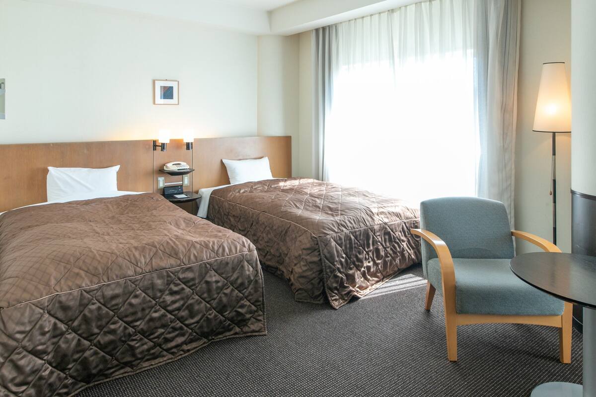 Ein Hotelzimmer mit zwei Betten, einem Sessel, einem Tisch, einer Lampe und einem Nachttisch.