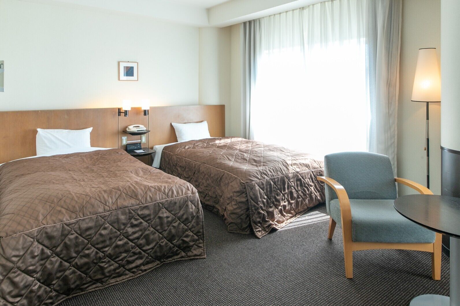 Habitació d'hotel amb dos llits, una taula, una cadira, una làmpada i una finestra amb cortines.