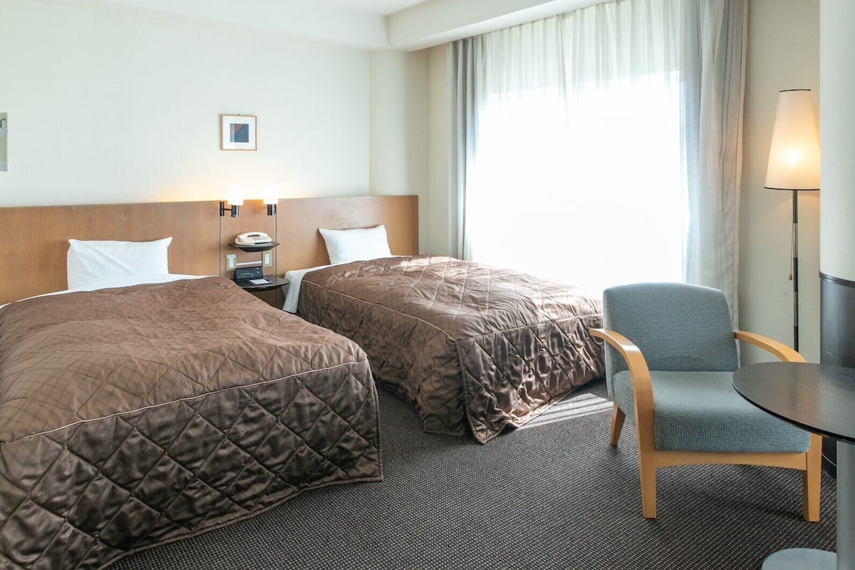 Ein Hotelzimmer mit zwei Betten, einem Schreibtisch, einem Stuhl, einer Lampe und einem Fenster mit Vorhängen.