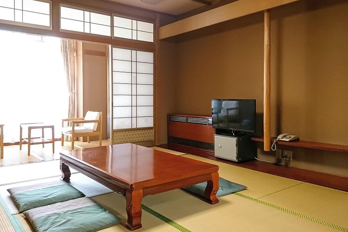 Une pièce de style japonais traditionnel, avec un sol recouvert de tatamis, une petite table basse en bois, un téléviseur et un petit meuble.