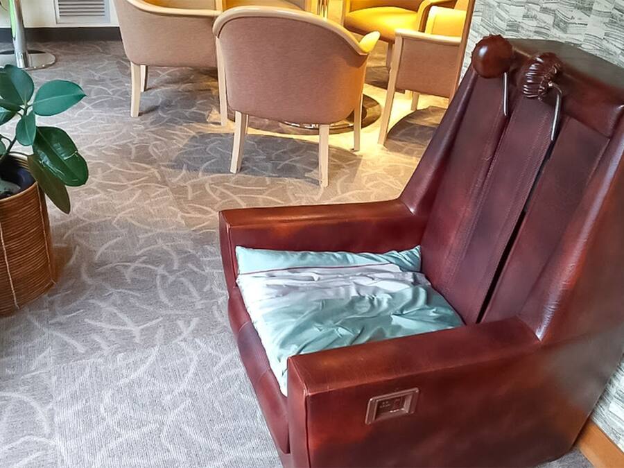 Ein brauner Ledersessel mit hellblauer Sitzkissen in einem Raum mit gemustertem Teppich, einer Korbpflanze und einem gelben Sofa im Hintergrund.