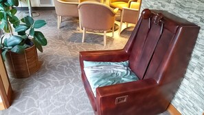 Ein brauner Ledersessel mit hellblauer Sitzkissen in einem Raum mit gemustertem Teppich, einer Korbpflanze und einem gelben Sofa im Hintergrund.
