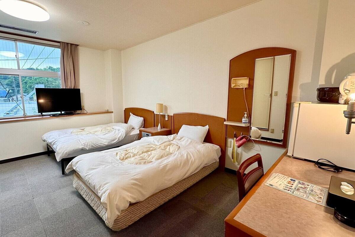 A hotel room with two beds, a television, a desk with a chair, a mirror, and a window with a view of the outside.