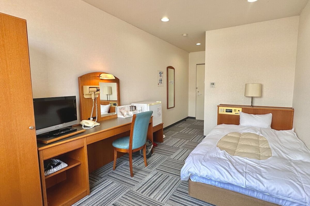 A hotel room with a bed, a desk with a television, a chair, and a bedside table with a lamp.