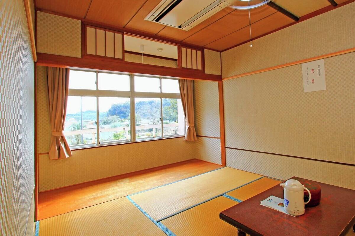 A traditional Japanese room with tatami flooring, a futon bed, a table with a kettle, and a view of a landscape.