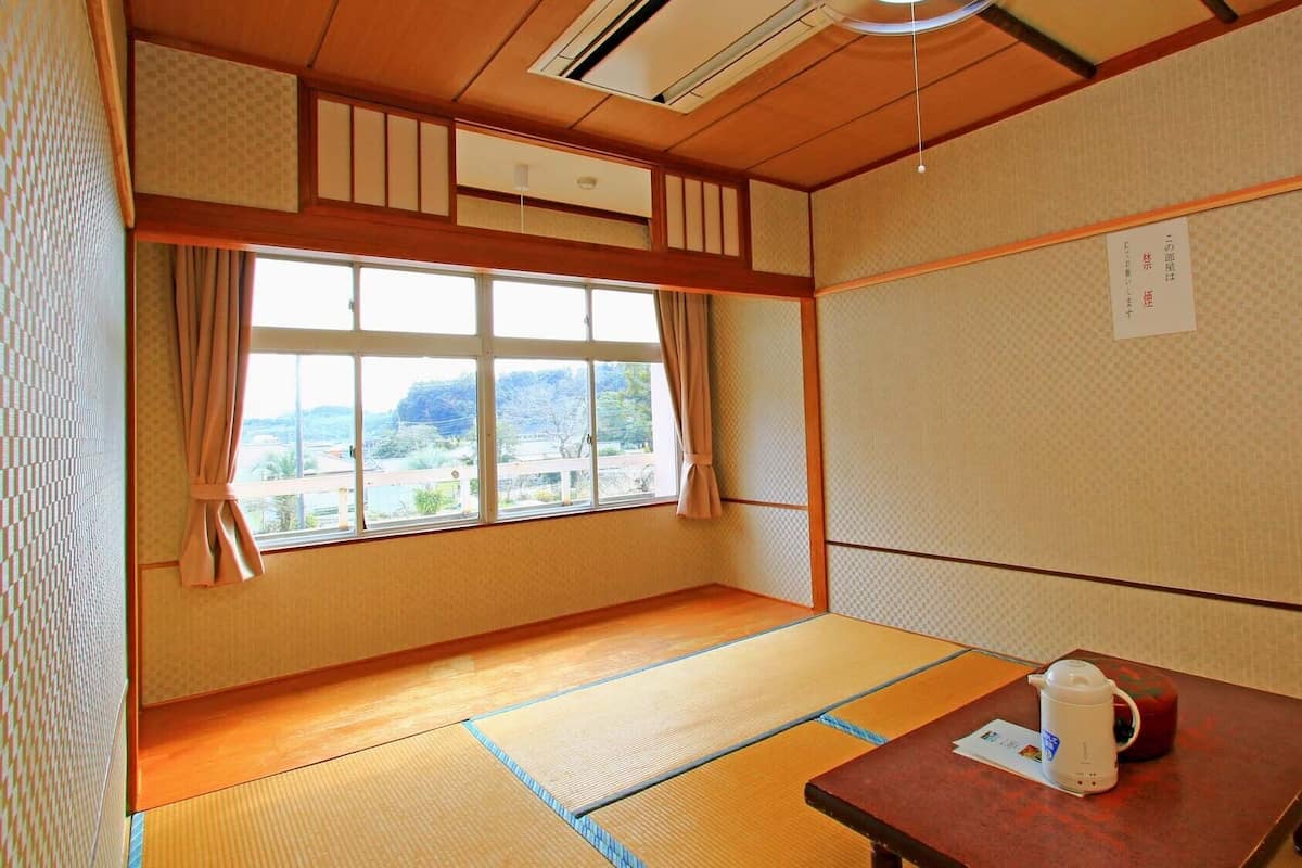 A traditional Japanese room with tatami flooring, a wooden table with a kettle, and a window with a view of a landscape.