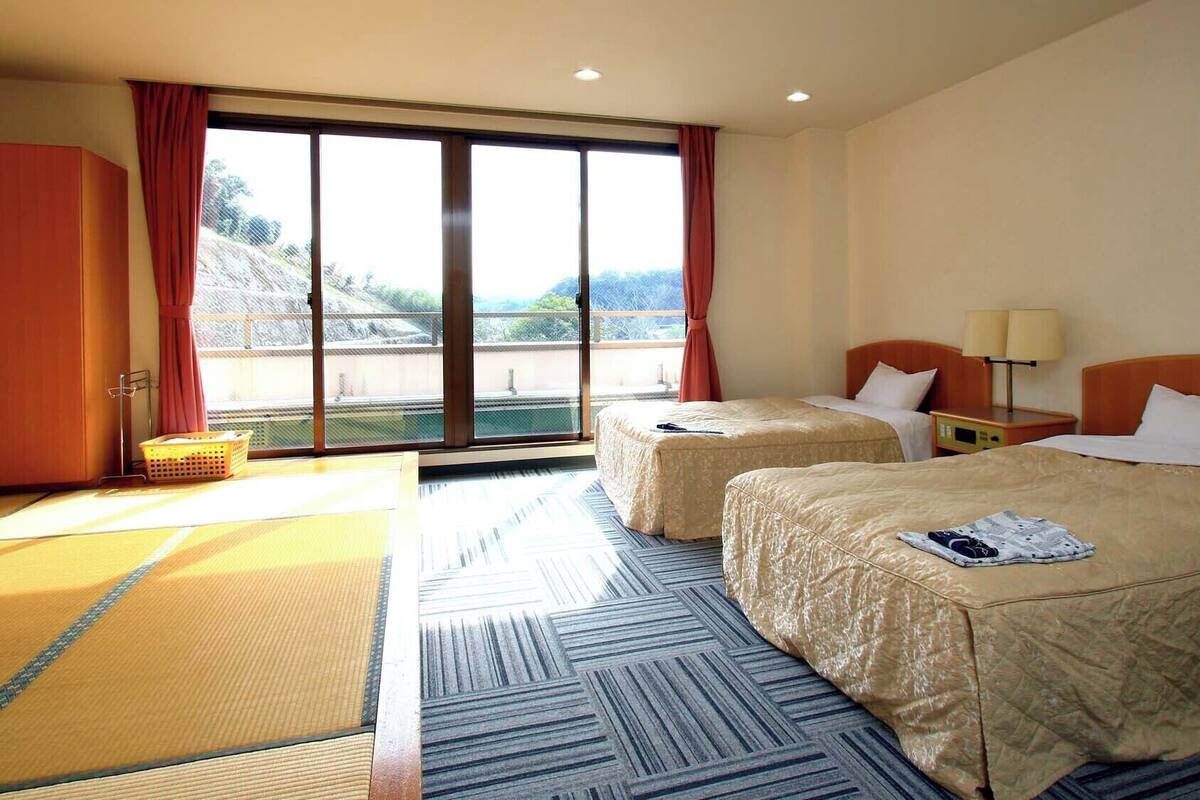 A hotel room with two beds, a desk, a chair, and a large window with a view of a mountainous landscape.
