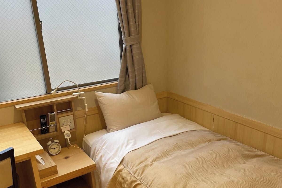 Ein kleines Zimmer mit einem Einzelbett, einem Holztisch, einem Nachttischtelefon und einem Fenster mit Vorhängen.