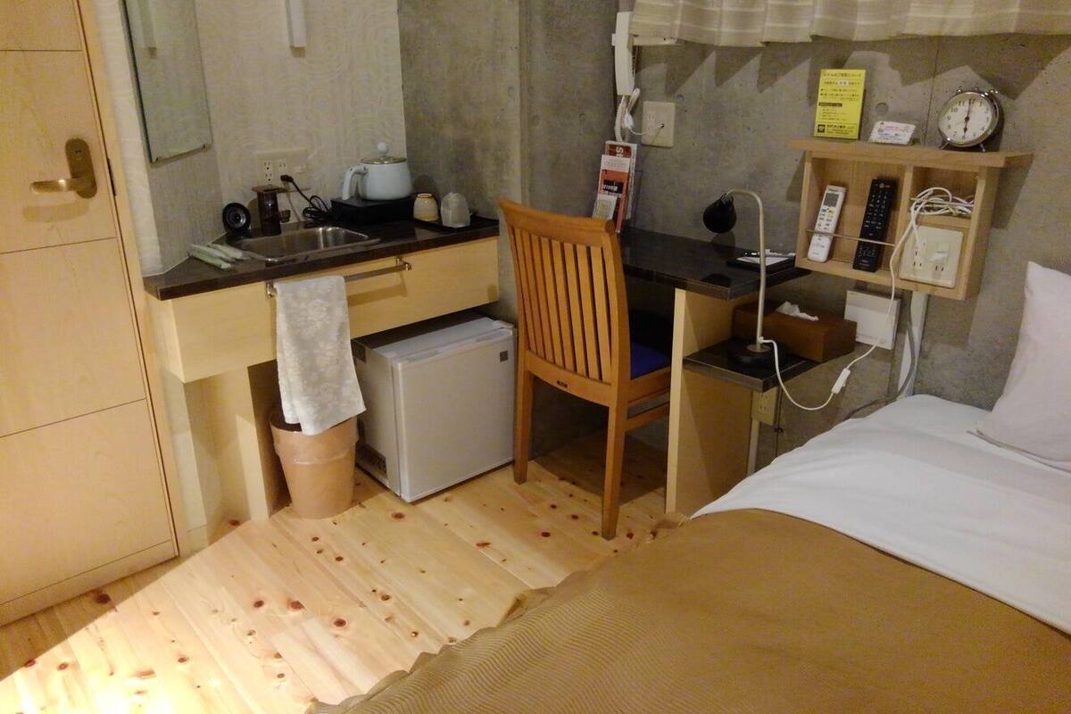 Ein kleines Hotelzimmer mit Küchenzeile, einem Einzelbett, einem Holzstuhl, einem Schreibtisch und einem Nachttisch mit Lampe.