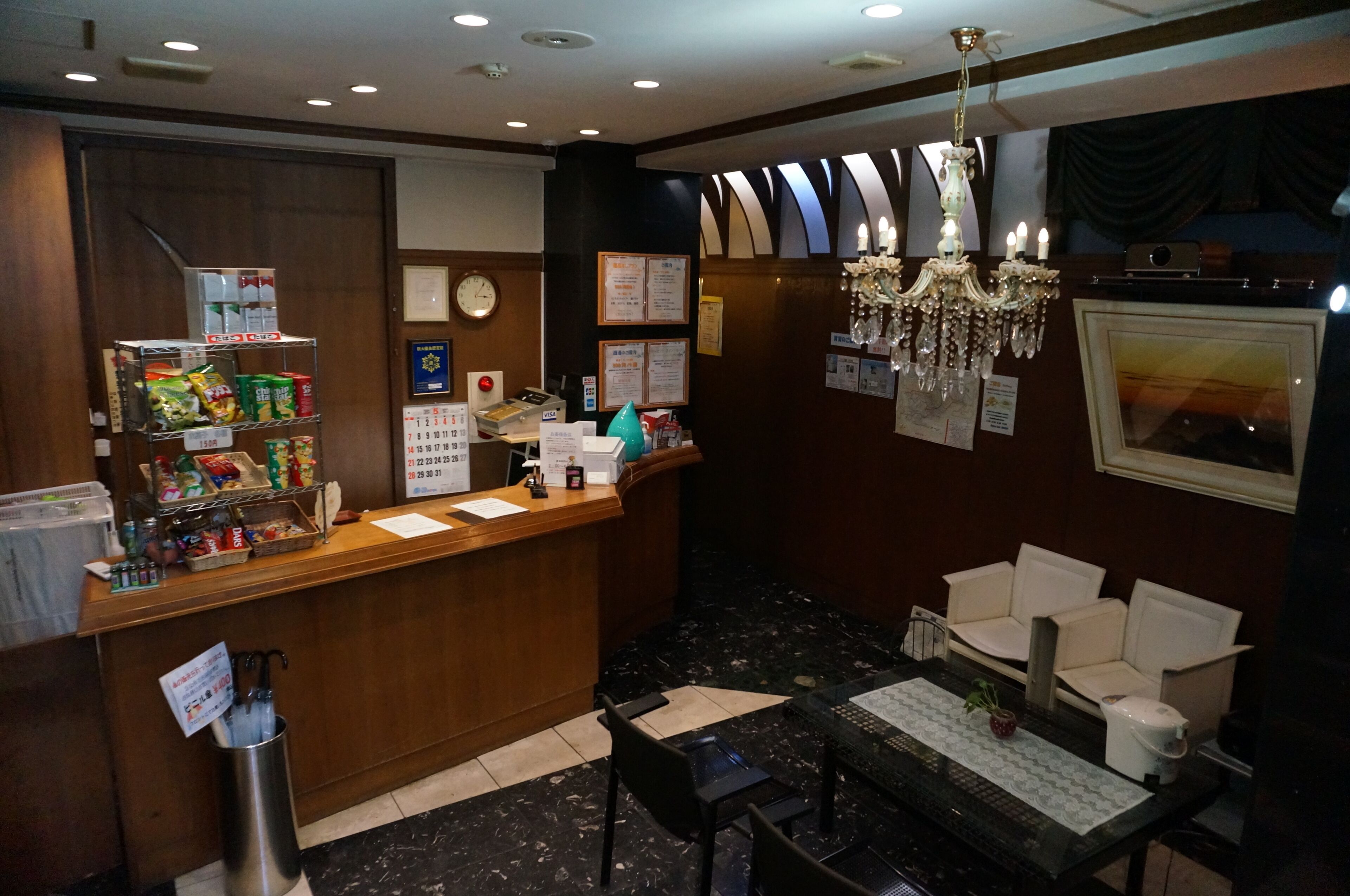 A hotel reception area with a wooden counter, a chandelier, a clock, and a seating area with chairs.