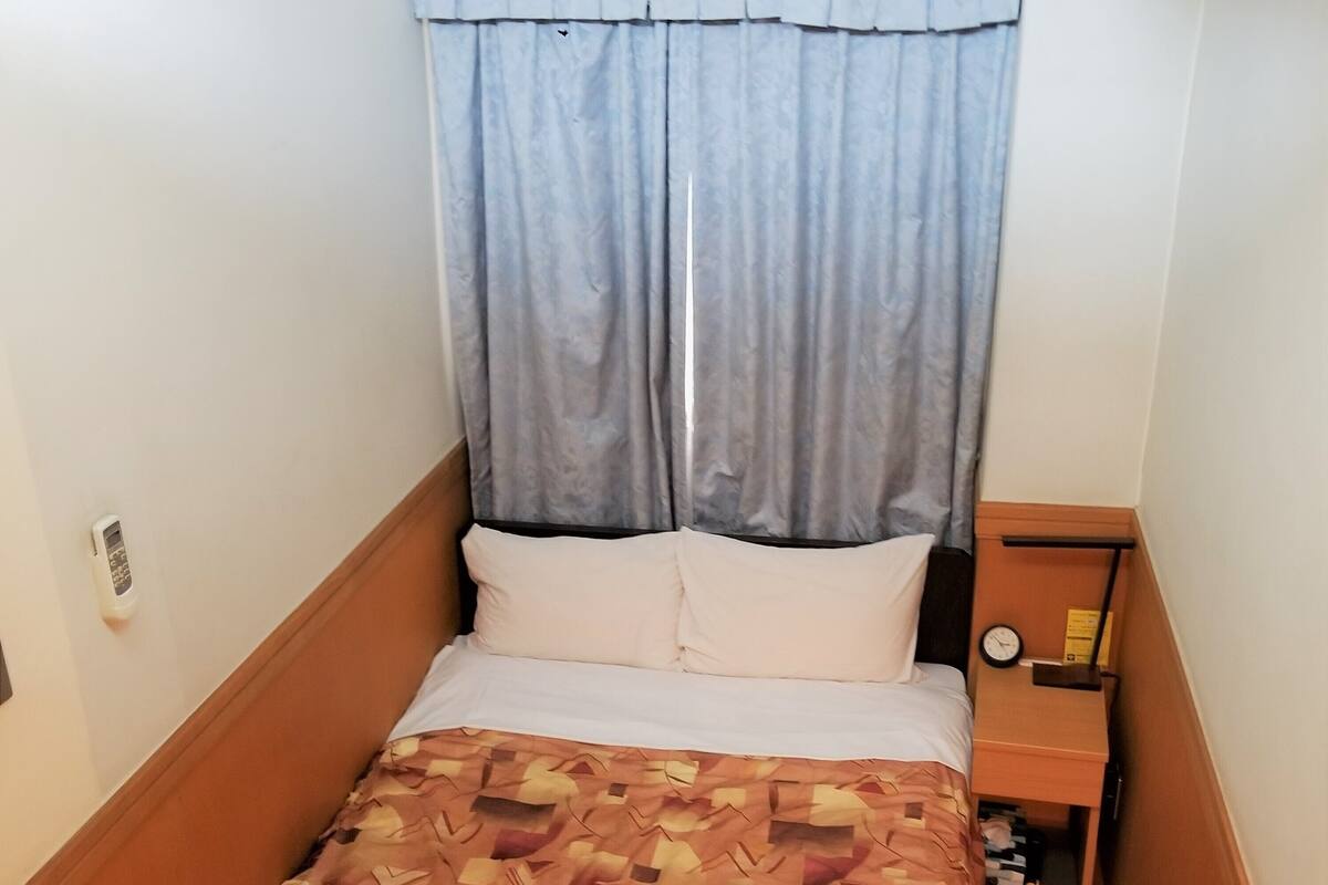 Ein kleines Zimmer mit einem Einzelbett, einer Bettdecke mit Muster, einem Nachttisch mit einer Uhr und einem Vorhang.
