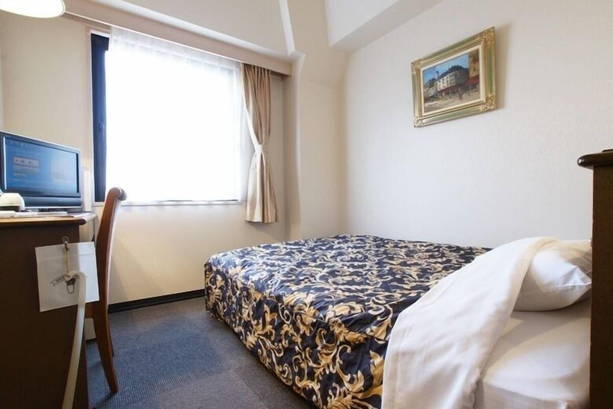 A hotel room with a bed, a desk with a computer, a chair, a window with curtains, and a framed picture on the wall.