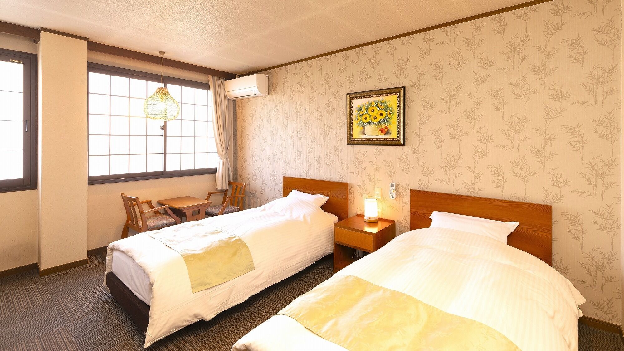 A hotel room with two beds, a desk, chairs, a window with curtains, a wall-mounted air conditioner, and a framed picture on the wall.