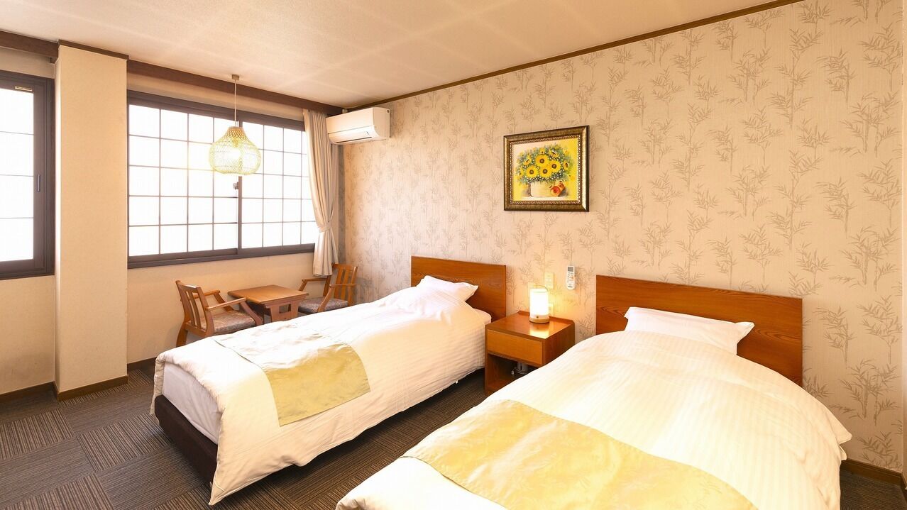 Ein Hotelzimmer mit zwei Betten, einem Schreibtisch, einem Stuhl, einem Fenster mit Vorhängen, einer Wand-Klimaanlage und einem gerahmten Bild an der Wand.