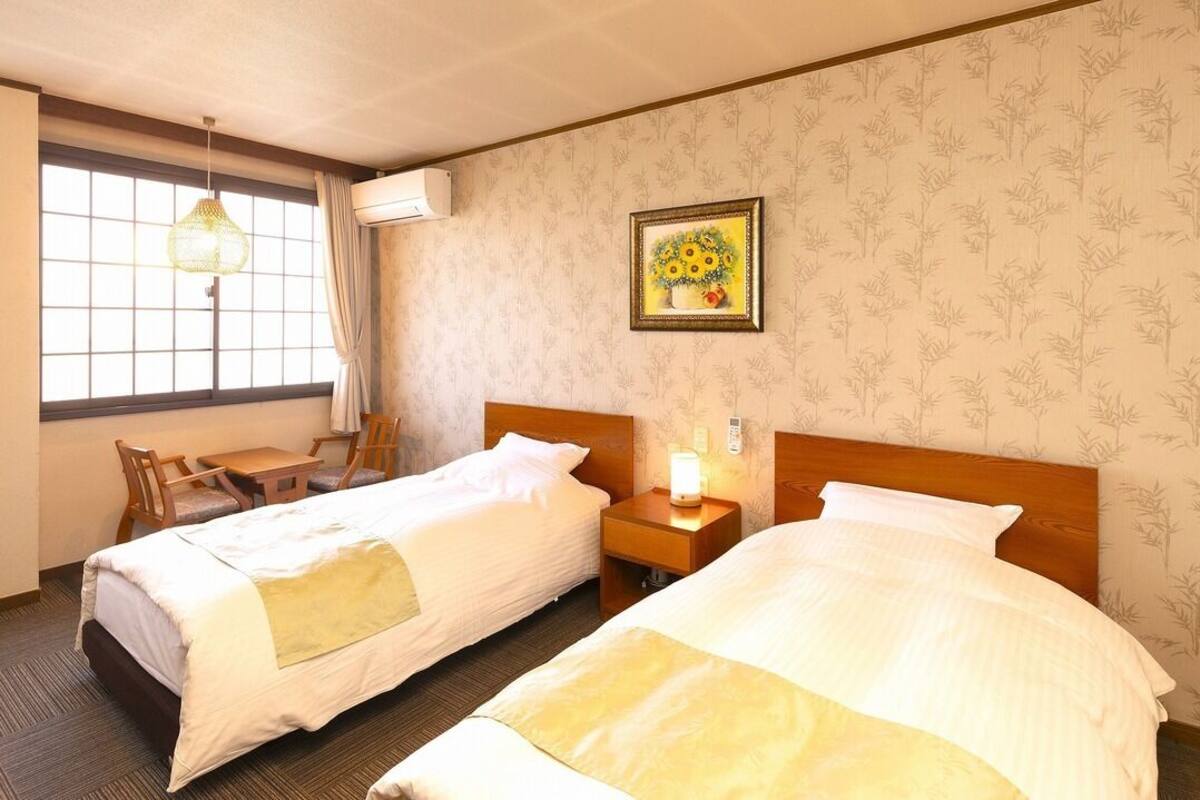 A hotel room with two beds, a desk, a chair, a window with curtains, a wall-mounted air conditioner, and a framed picture on the wall.