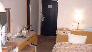 A hotel room with a bed, a desk with a chair, a mirror, a bedside table with a lamp, and a door. - Imabari Plaza Hotel (Imabari)