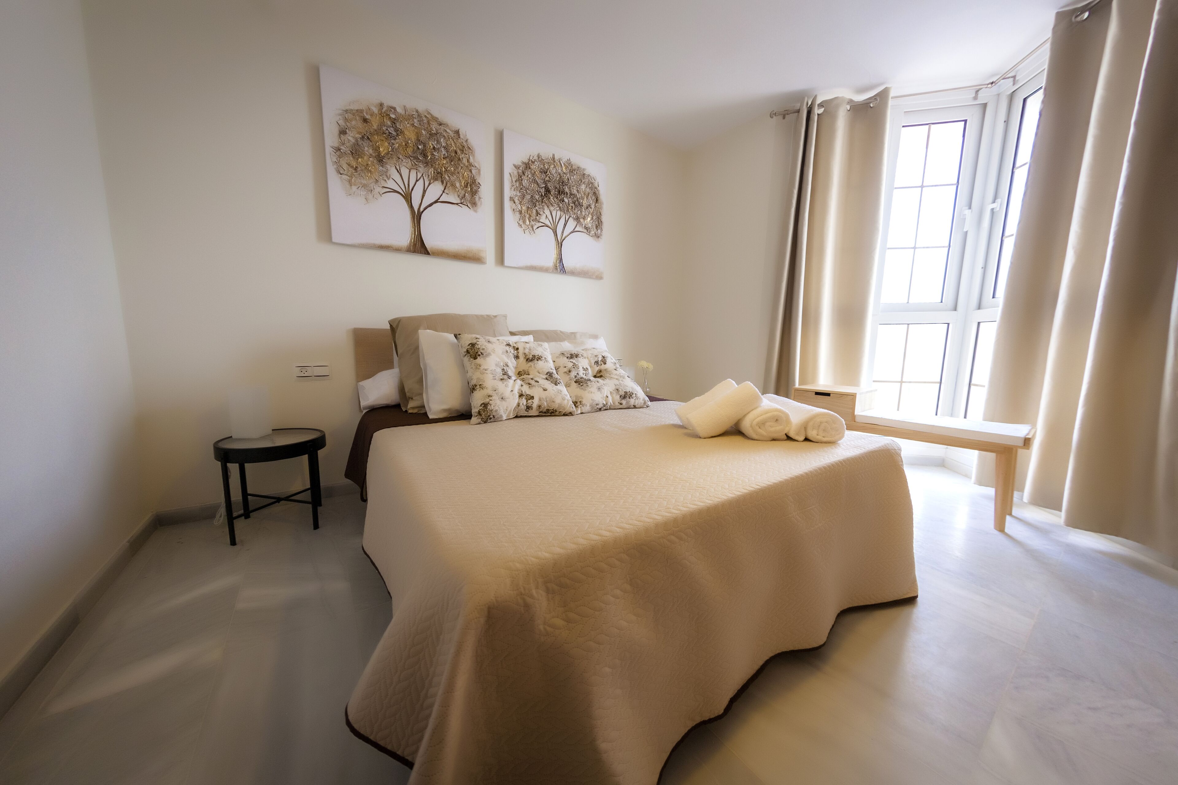 Un dormitorio con una cama grande, una mesita de noche, un taburete pequeño y dos cuadros colgados en la pared.