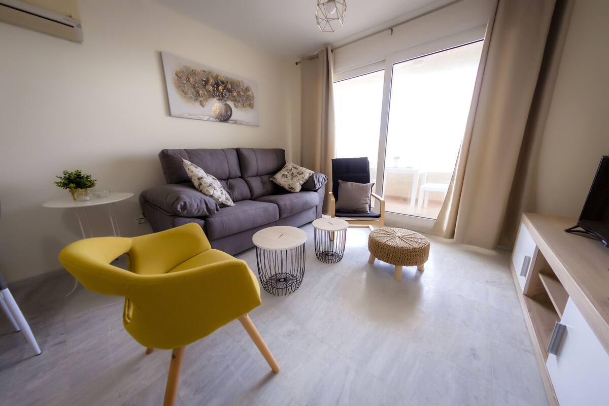 Un salon moderne avec un canapé gris, des fauteuils jaunes, une petite table ronde et un téléviseur posé sur un meuble en bois.