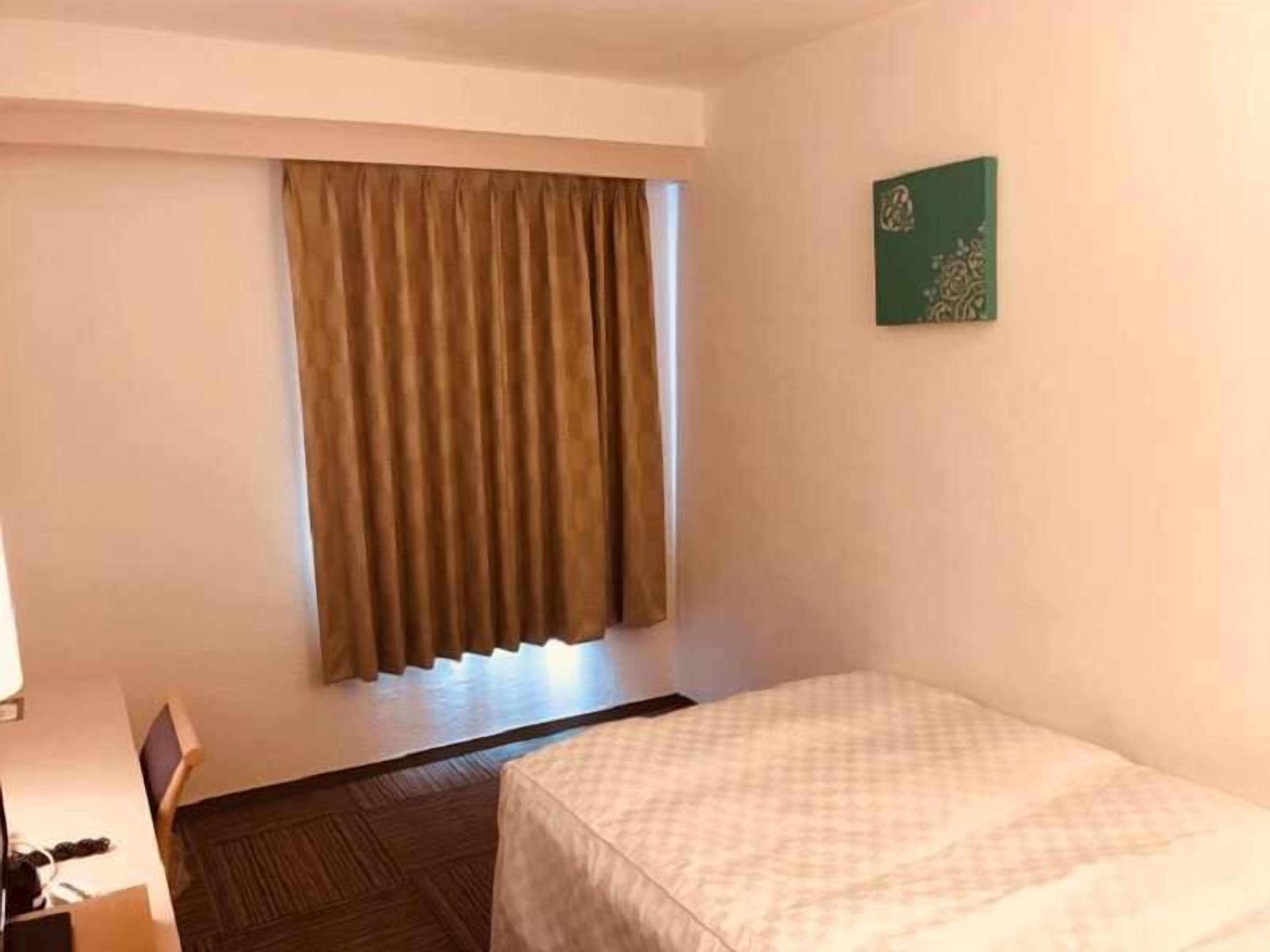 A hotel room with a bed, a window with curtains, a small table, and a wall-mounted artwork.