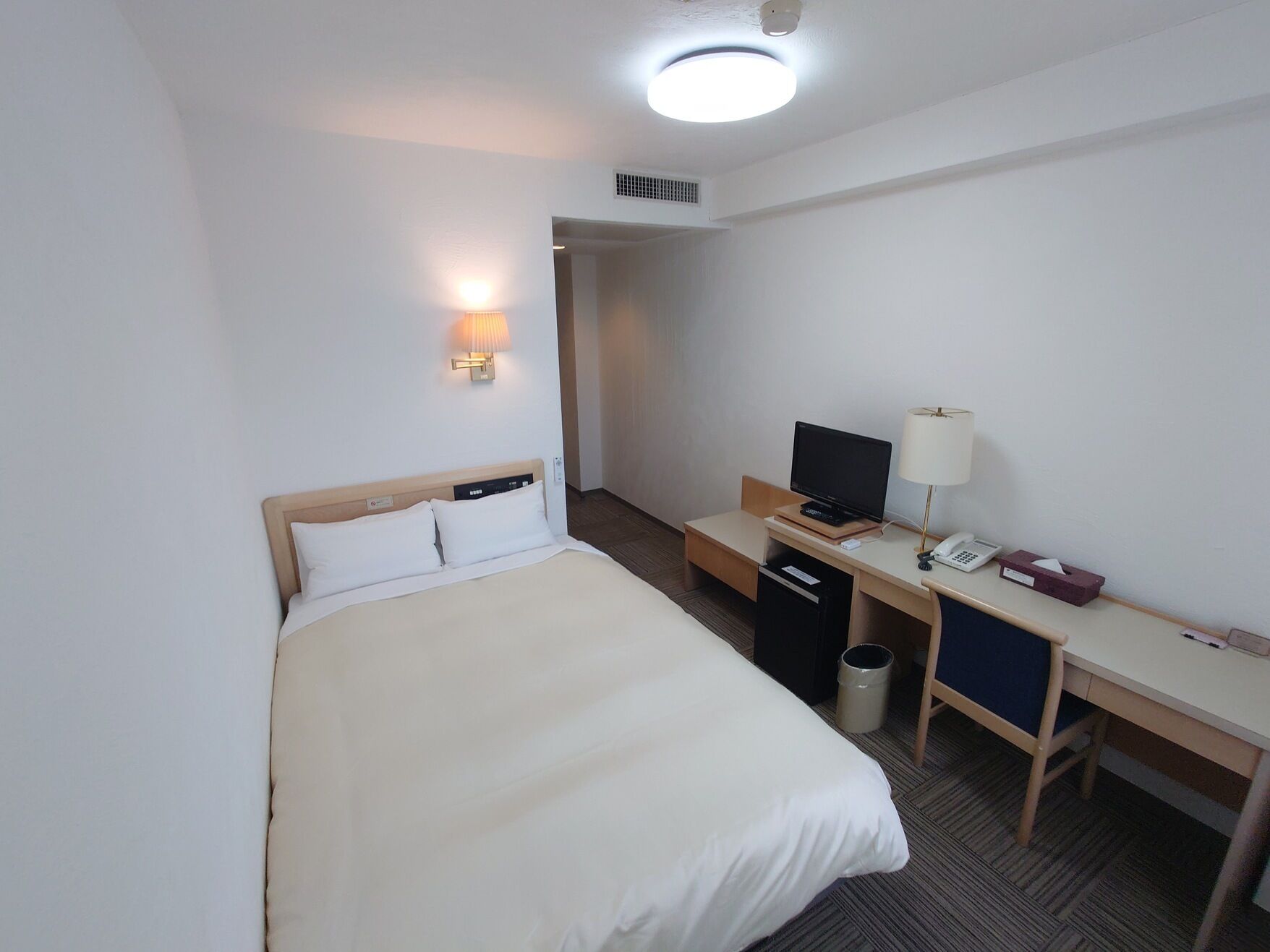 A hotel room with a bed, desk, chair, TV, lamp, and a trash can.
