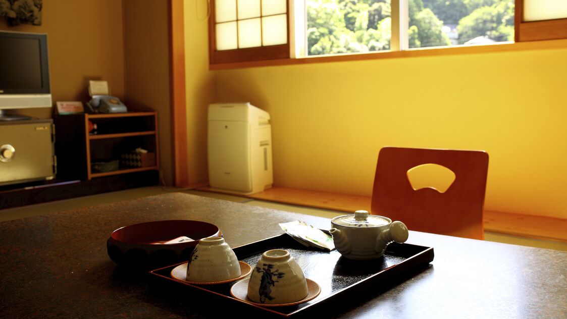 Une pièce de style japonais traditionnel, avec un sol recouvert de tatamis, une petite table sur laquelle est disposée une sélection d’ustensiles à thé, un téléviseur et un petit réfrigérateur.
