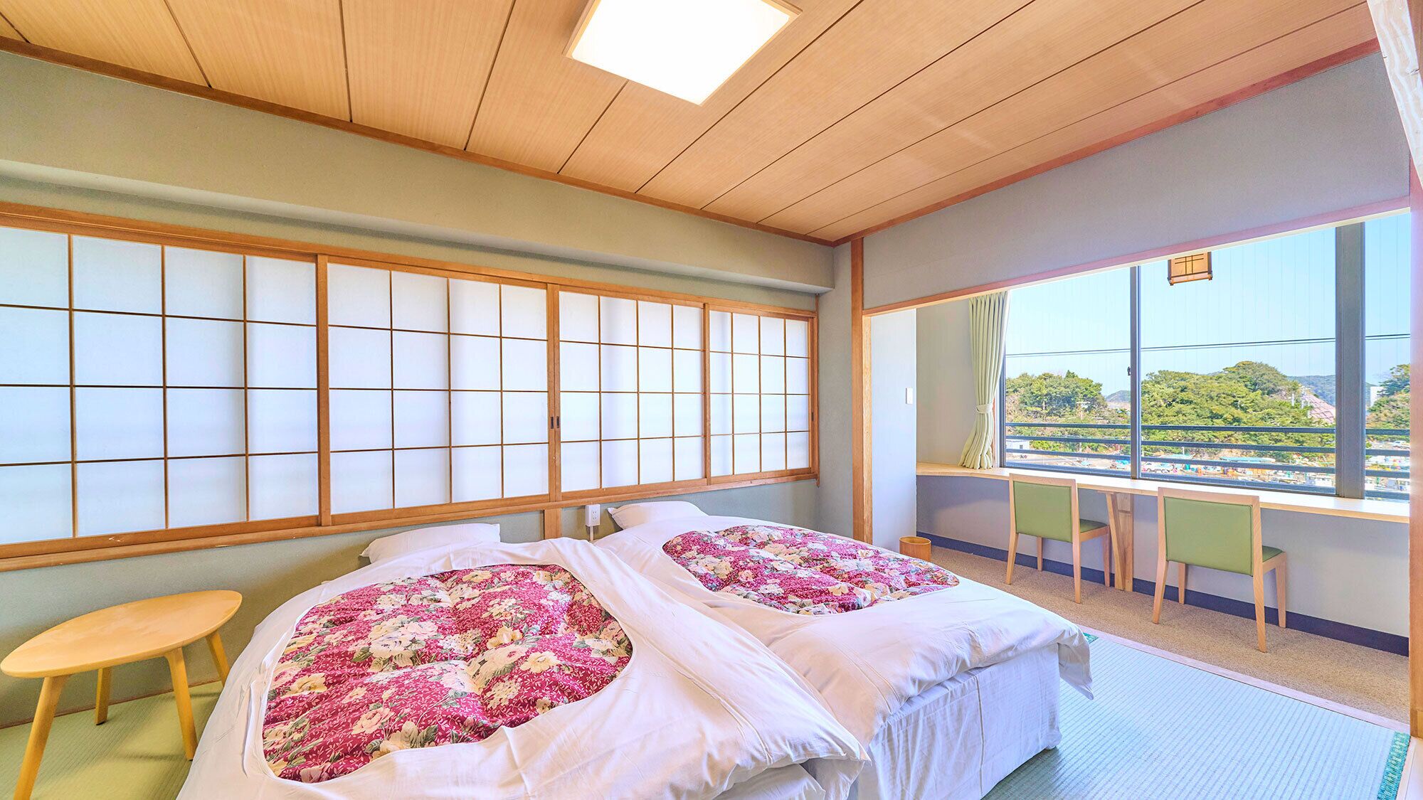 A room with tatami flooring, a traditional Japanese sliding door, a bed with floral bedding, a desk with a chair, and a view of greenery outside.