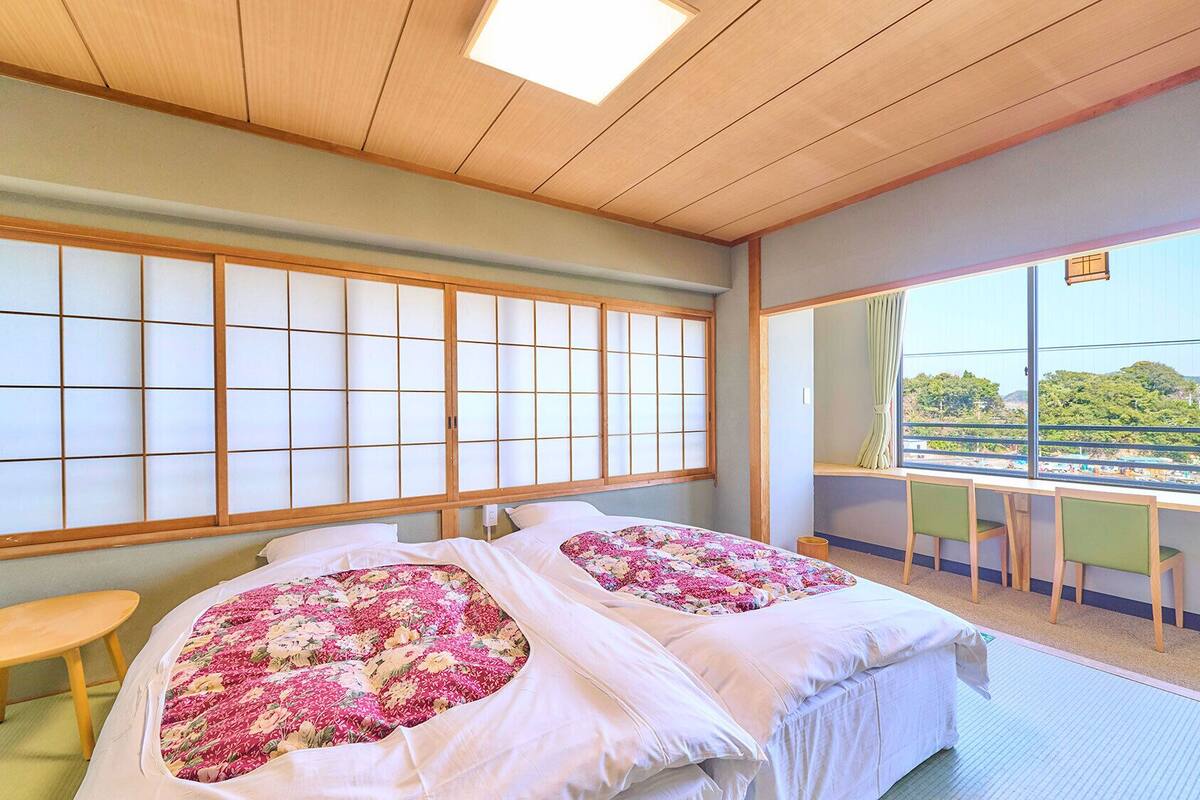 Ein Zimmer mit Tatami-Boden, einer traditionellen japanischen Schiebetür, einem Bett mit floralem Bettzeug, einem Schreibtisch mit Stuhl und Blick auf Grünflächen außerhalb.