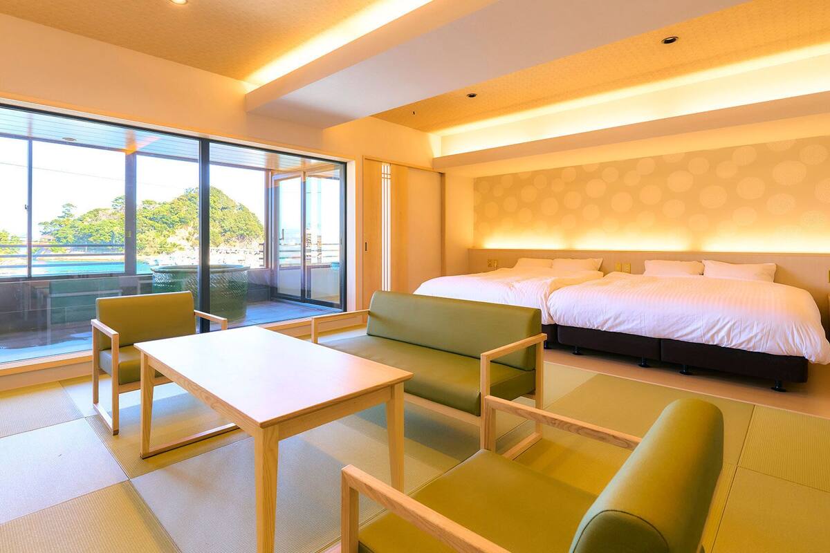Ein Hotelzimmer mit einem grossen Bett, einer Sitzecke mit Sesseln und einem Tisch sowie Blick auf einen Strand durch die Fenster.