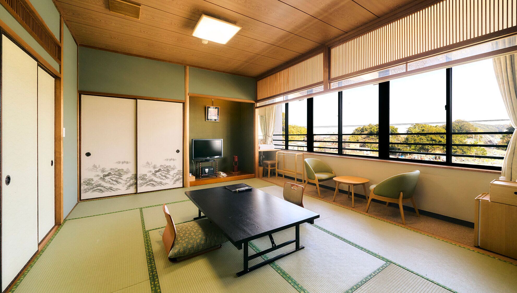 A traditional Japanese room with tatami flooring, a low table, chairs, a TV, and a view through large windows.