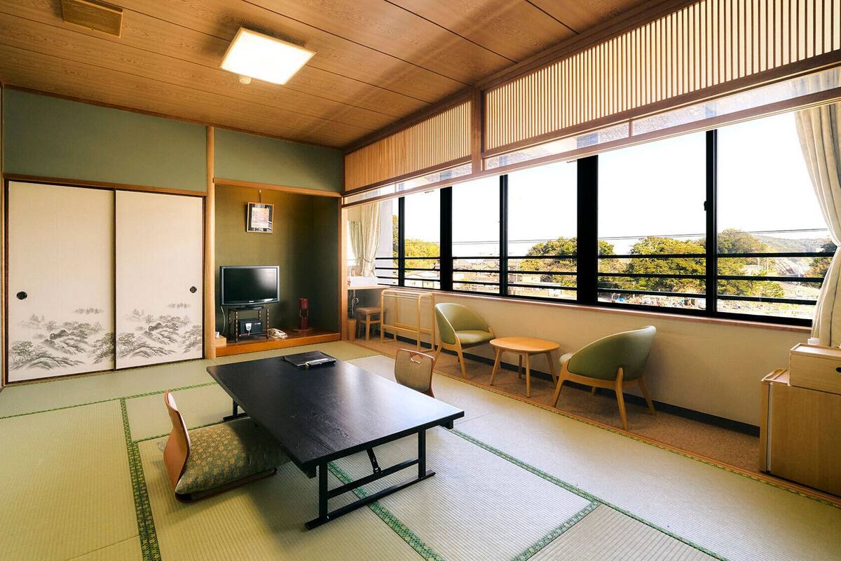 Ein traditioneller japanischer Raum mit Tatami-Boden, einem niedrigen Tisch, Stühlen, einem Fernseher und Blick durch große Fenster.