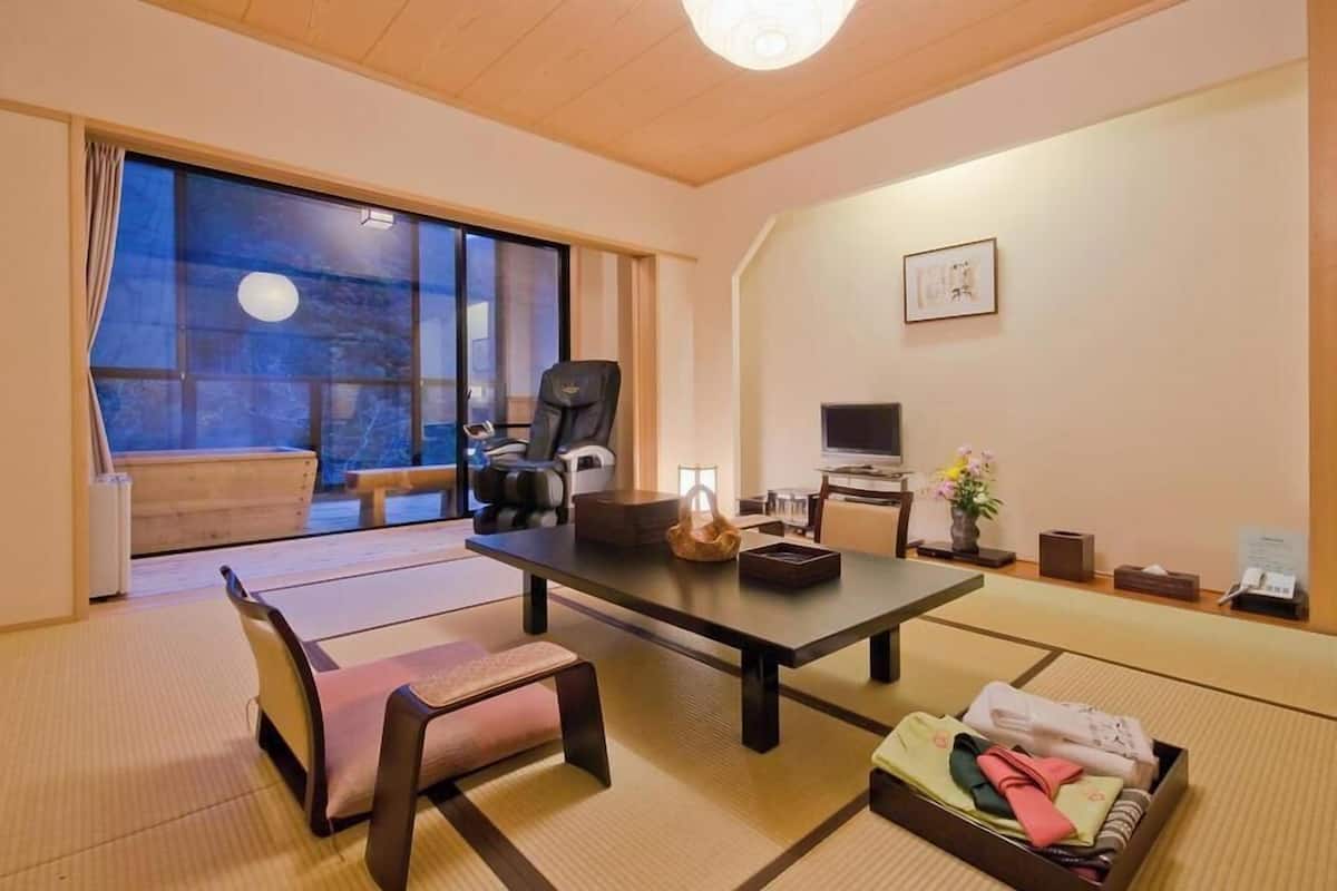 A traditional Japanese room with tatami flooring, a low table, a cushioned bench, a black chair, a television, and a sliding door leading to an outdoor area.