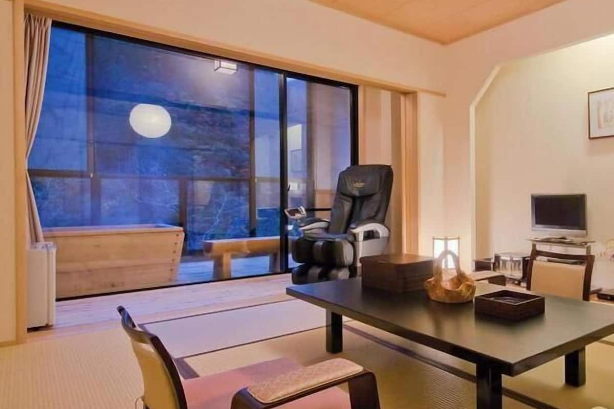 A room with tatami mats, a low table, a chair, a sofa, a television, and a view of the outdoors.