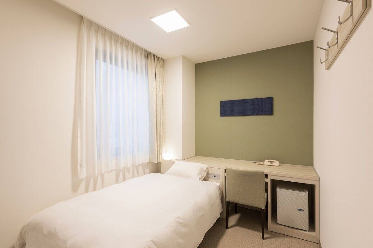 Una habitación de hotel con cama, escritorio, silla, mueble pequeño y ventana con cortinas.