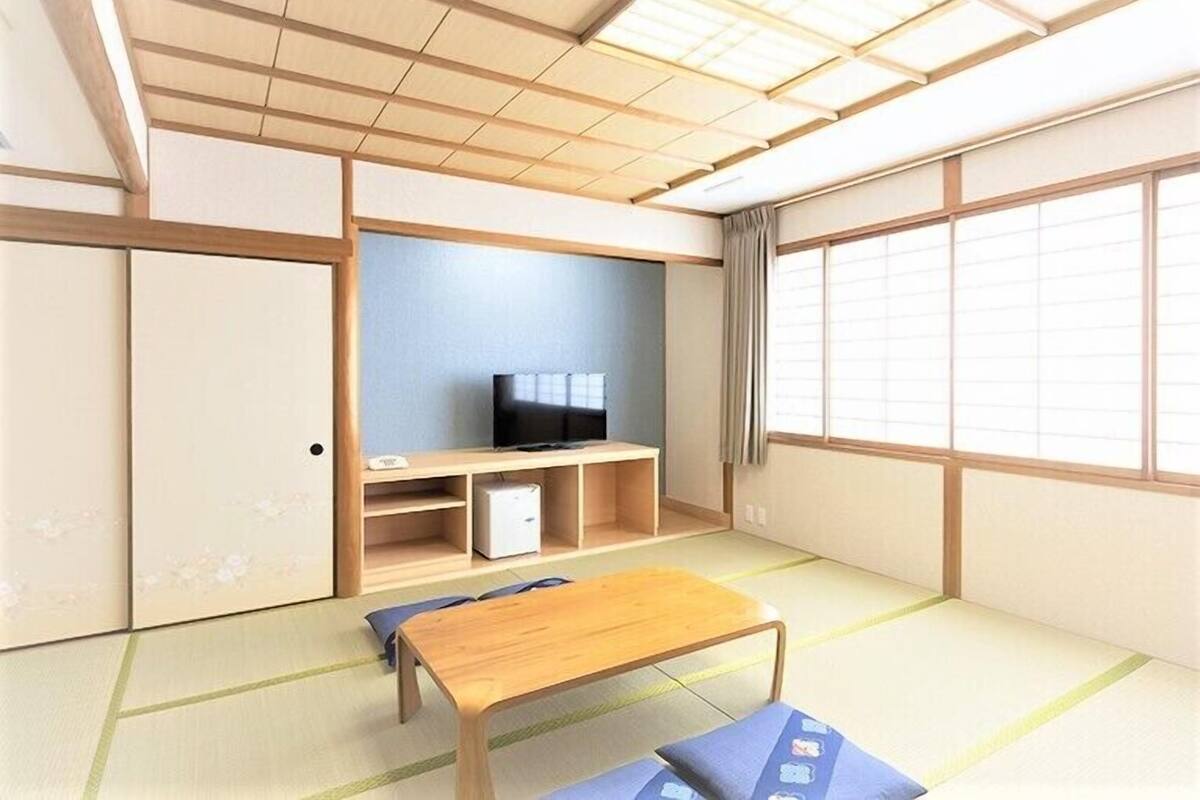 Una habitación con suelo de tatami, un televisor en una estantería, una mesa de madera y puertas correderas.