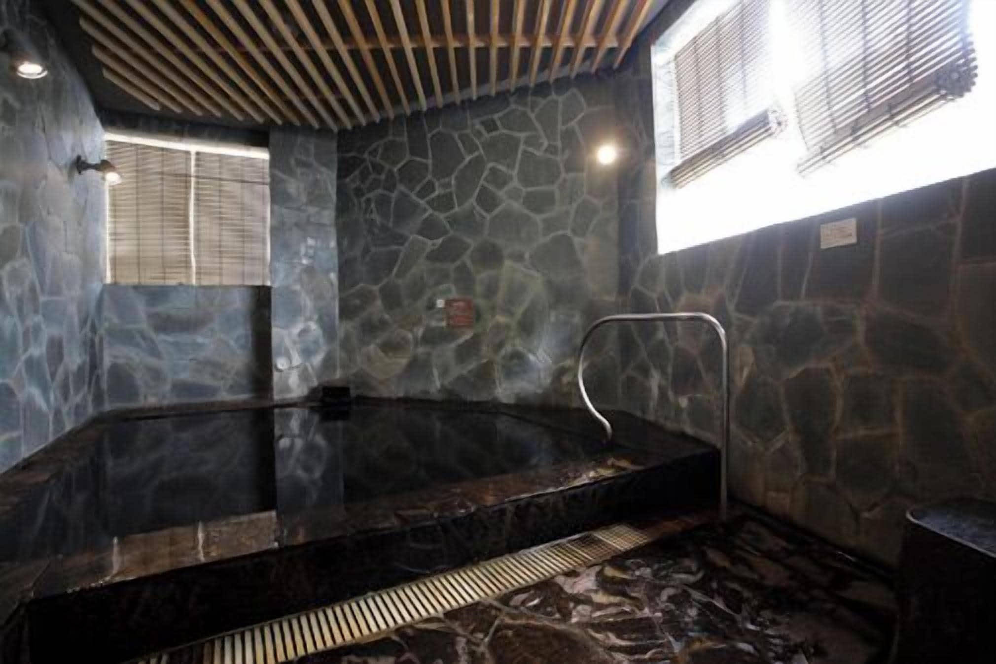 A stone-walled hot tub with a metal ladder, a window with blinds, and a ceiling with wooden slats.