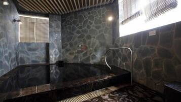 A stone-walled hot tub with a metal ladder, a window with blinds, and a ceiling with wooden slats.