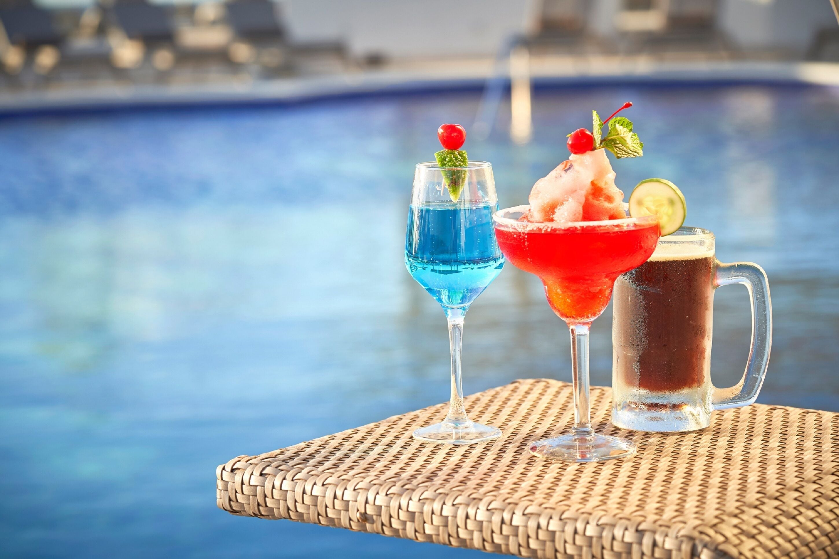 A poolside setting with a wicker table holding two drinks, one in a glass and one in a mug, with a pool in the background.