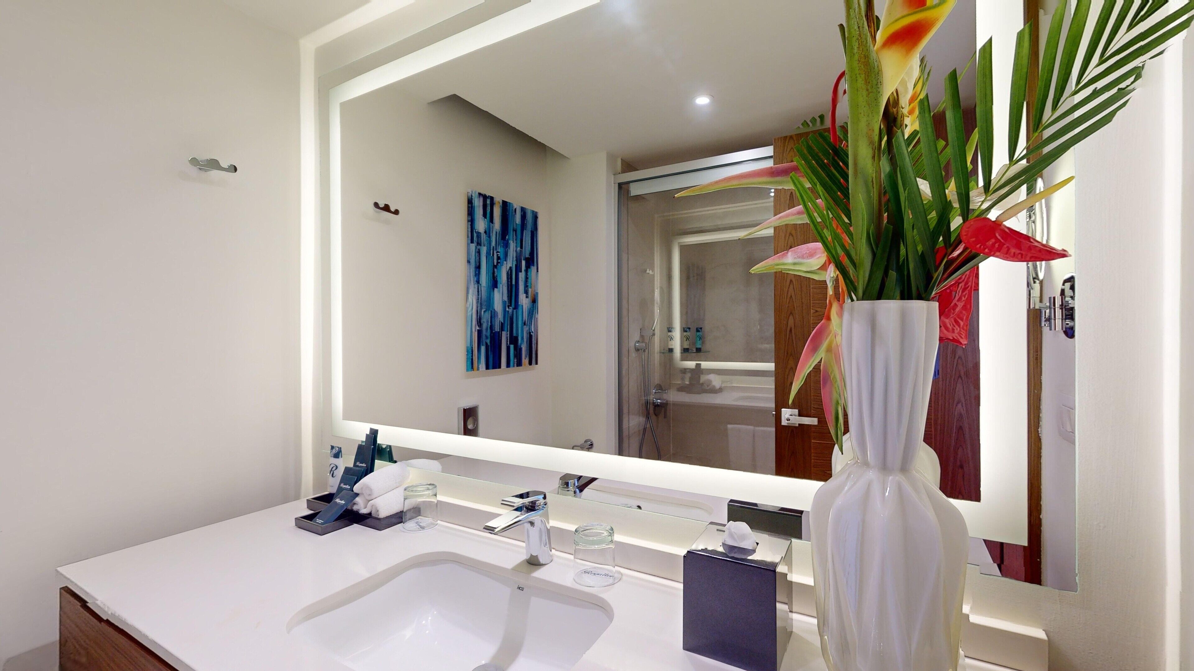 A modern bathroom with a white sink, a large mirror, and a vase with tropical flowers.
