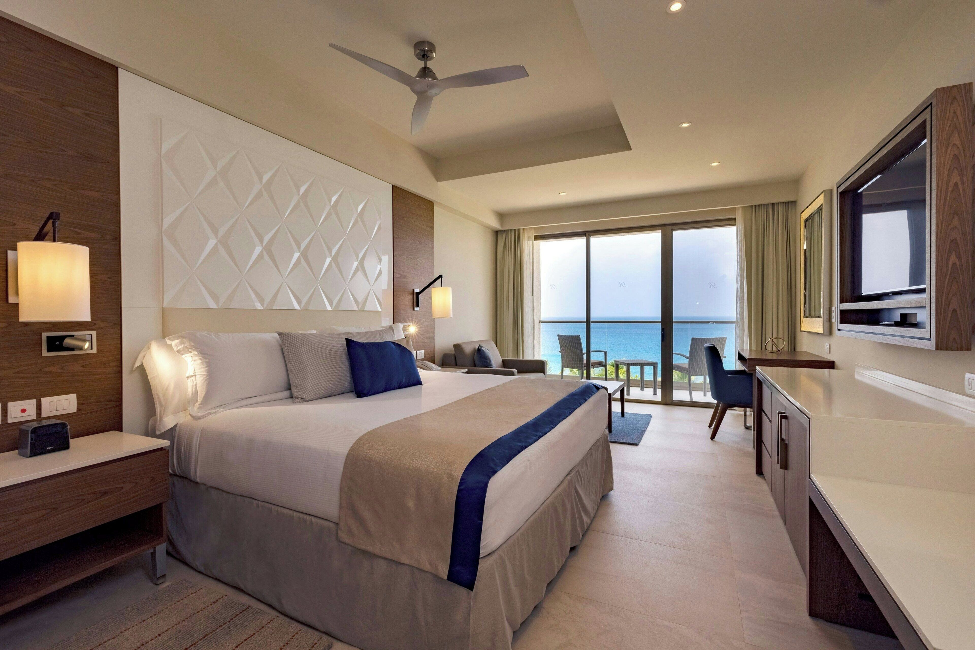 A modern hotel room with a large bed, a desk, a chair, a ceiling fan, and a view of the sea through the balcony.