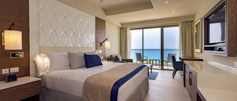 A modern hotel room with a large bed, a desk, a chair, a ceiling fan, and a view of the sea through the balcony.