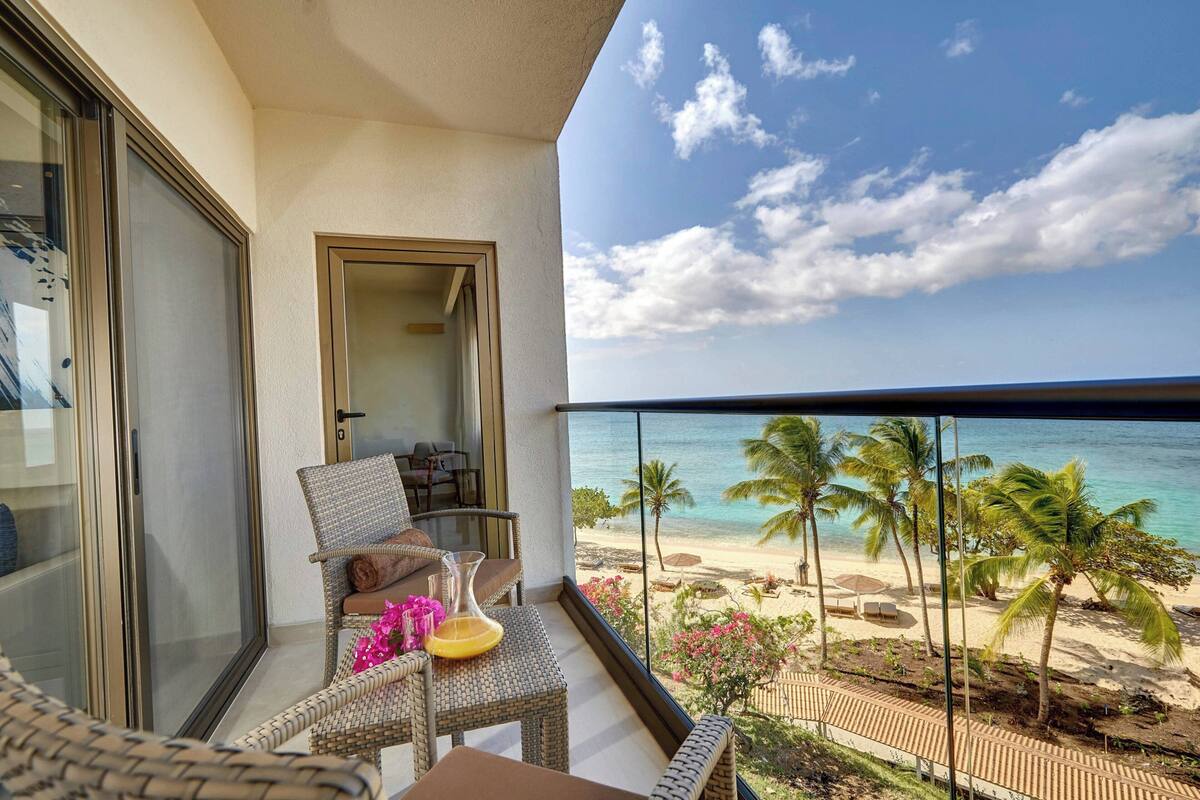 A balcony with wicker furniture, a glass door, and a view of a beach with palm trees and the ocean.