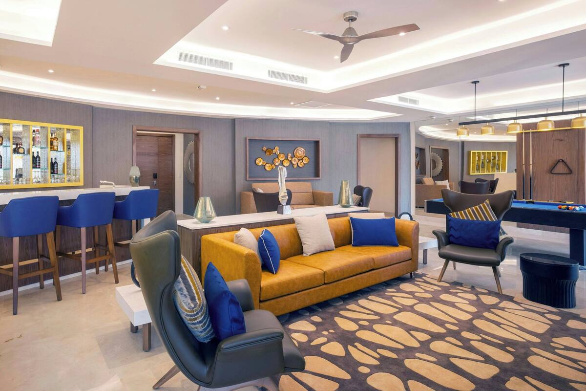 A modern living room with a yellow sofa, blue chairs, a pool table, and a bar area.