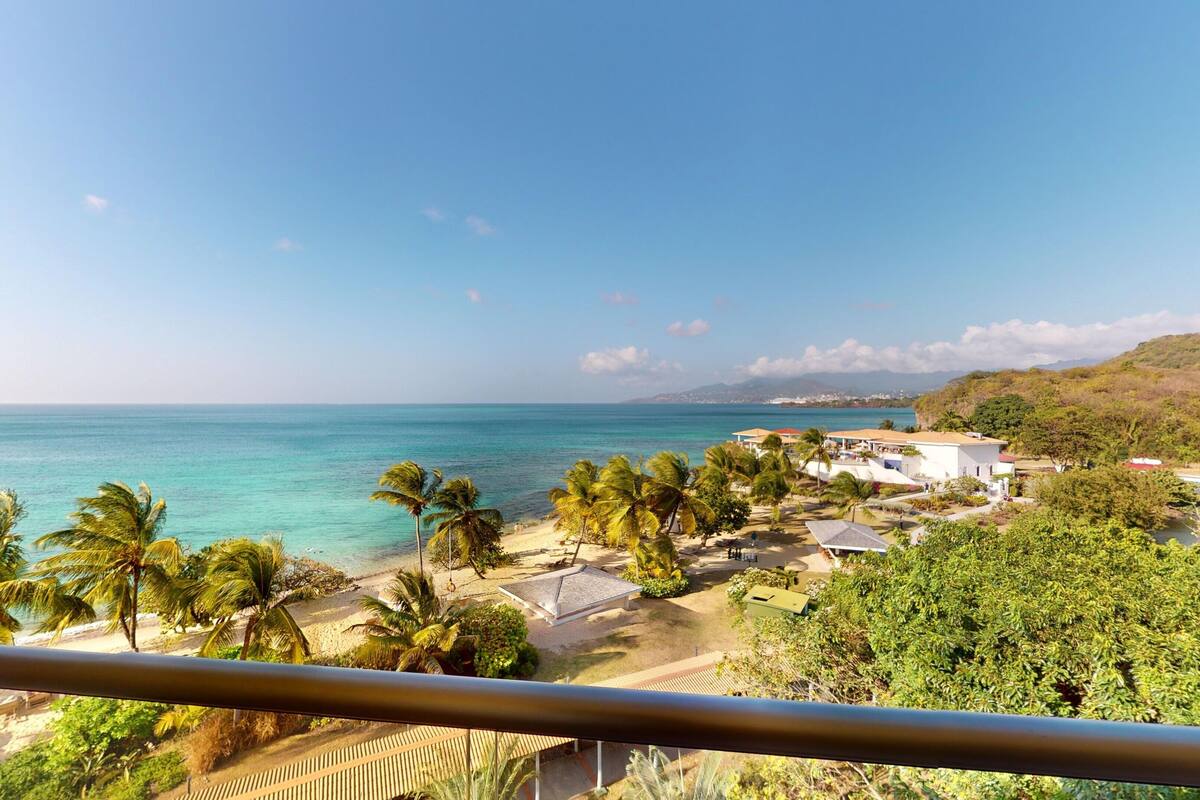 A coastal view with a beach, palm trees, and a white building with a red roof.