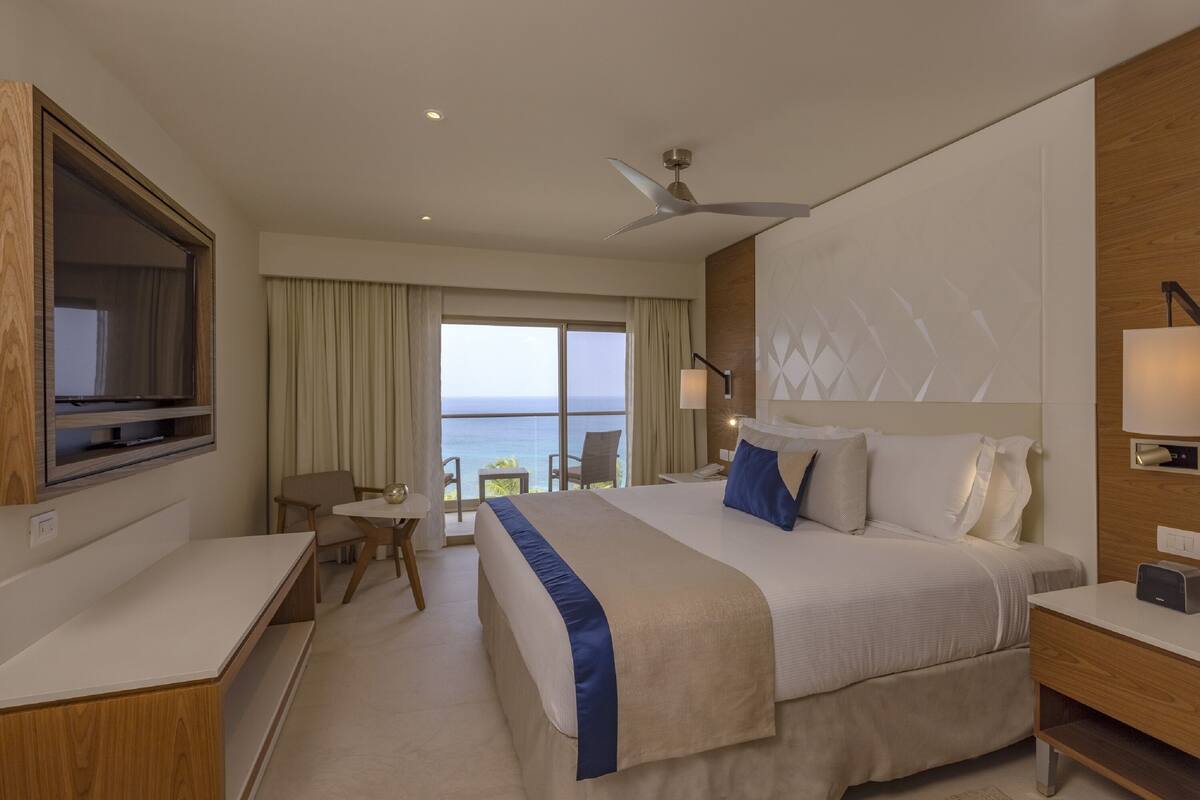 A hotel room with a large bed, a desk, a chair, a TV, and a balcony with a view of the sea.
