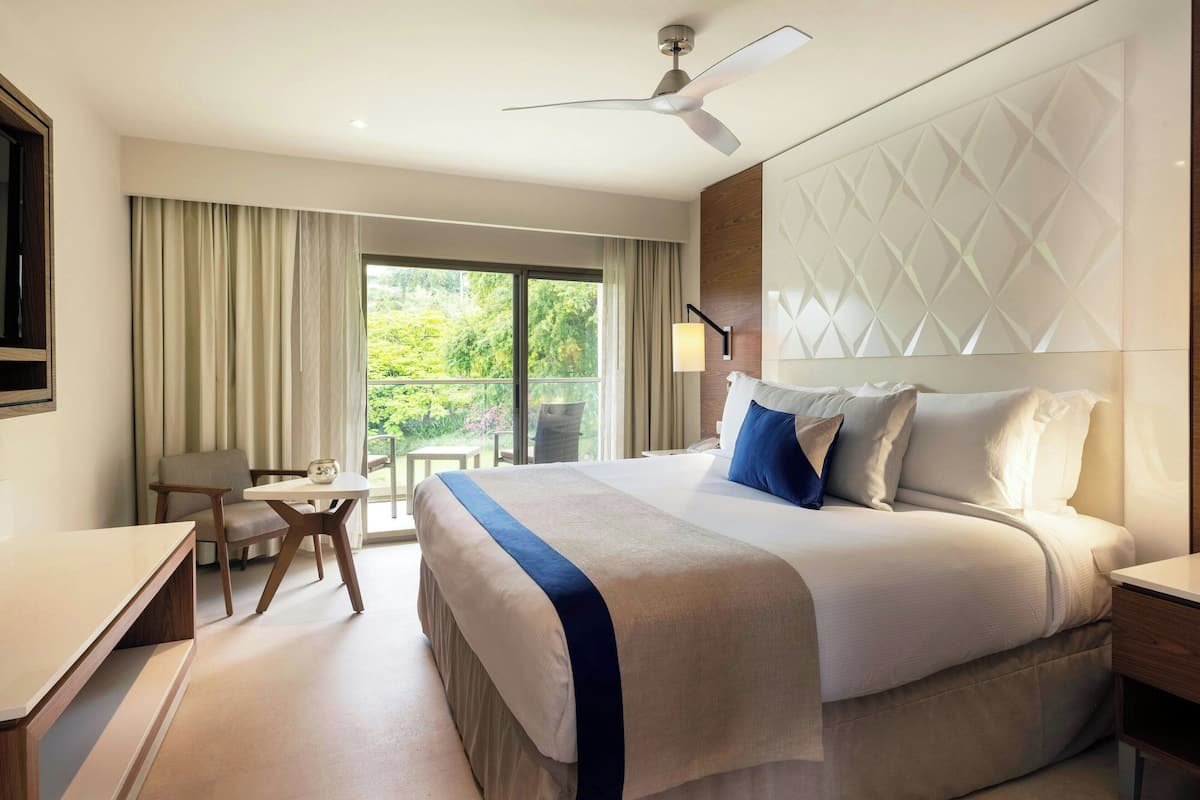 A hotel room with a large bed, a desk, a chair, and a view of greenery through the window.